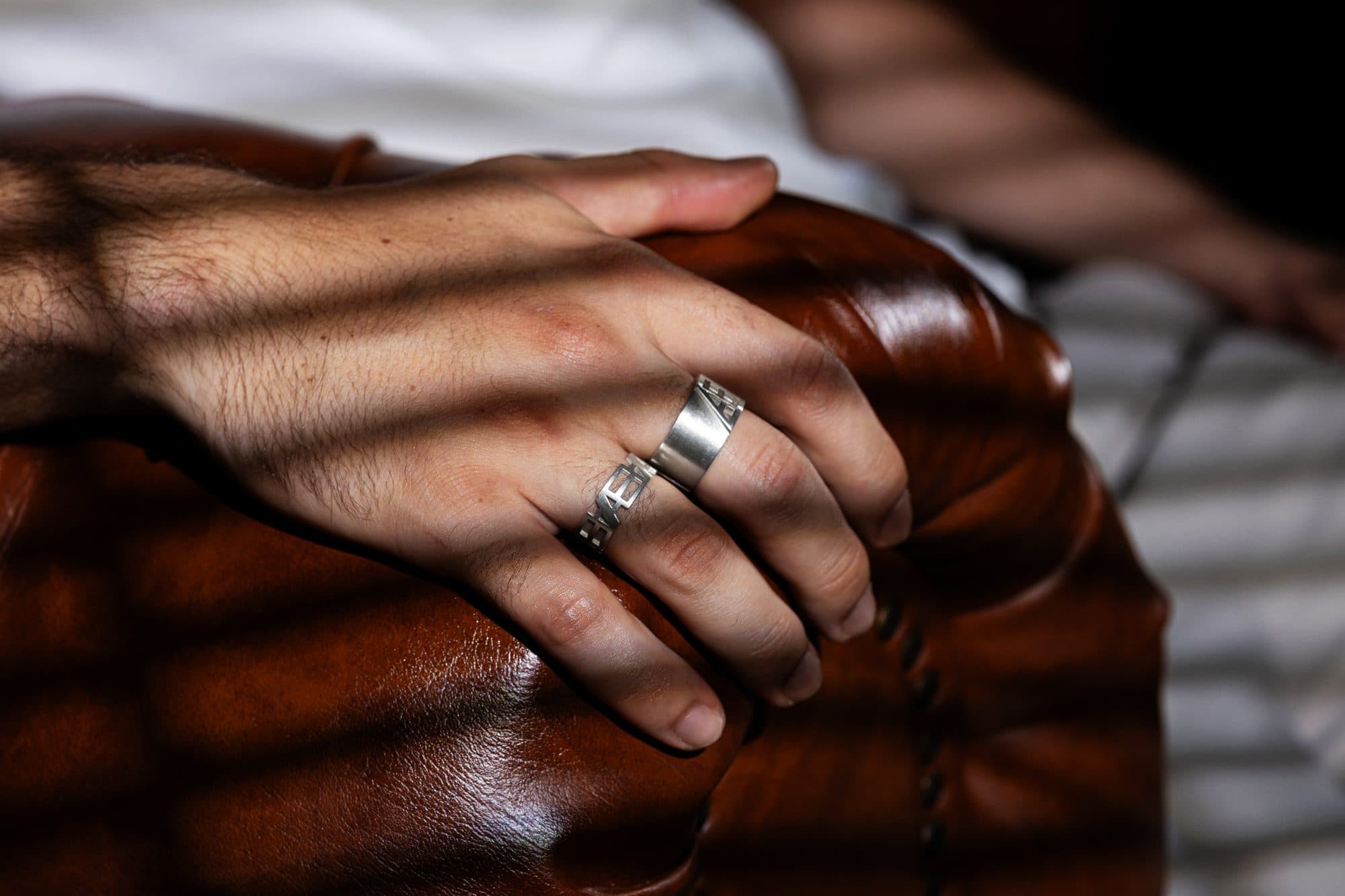 Hand wearing ÆTHERNO rings resting on a leather armchair in striped light